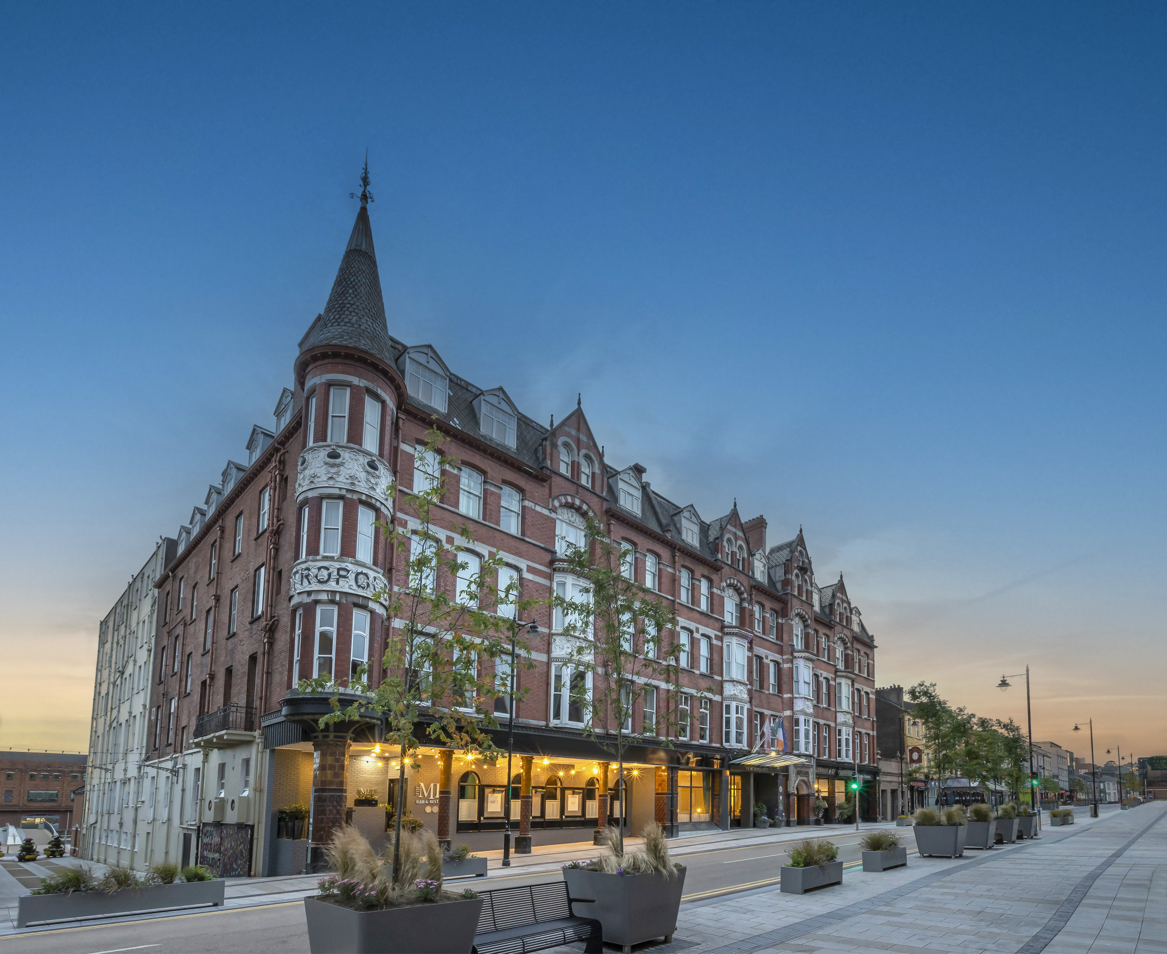 The red-brick exterior of The Metropole Hotel on MacCurtain Street in Cork&rsquo;s Victorian Quarter at dusk, with its turret, lit entrance and wide city pavement in view.
